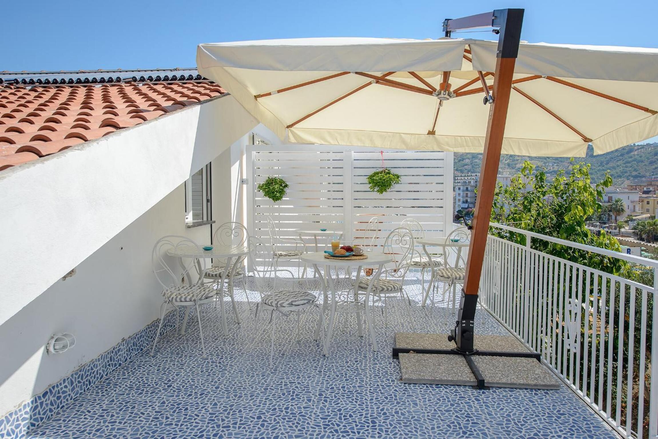 Una terraza con una mesa y sillas blancas, un paraguas color beige y una valla blanca con follaje.