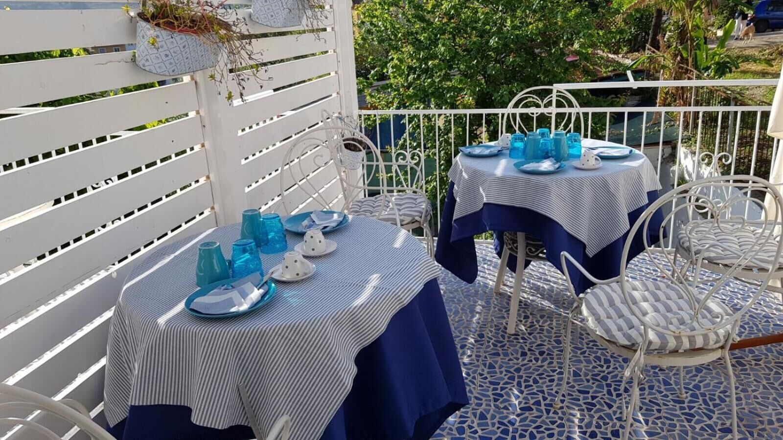 Dos mesas al aire libre dispuestas con manteles azules y cristalería azul a juego, rodeadas de sillas de metal blanco con cojines a rayas.