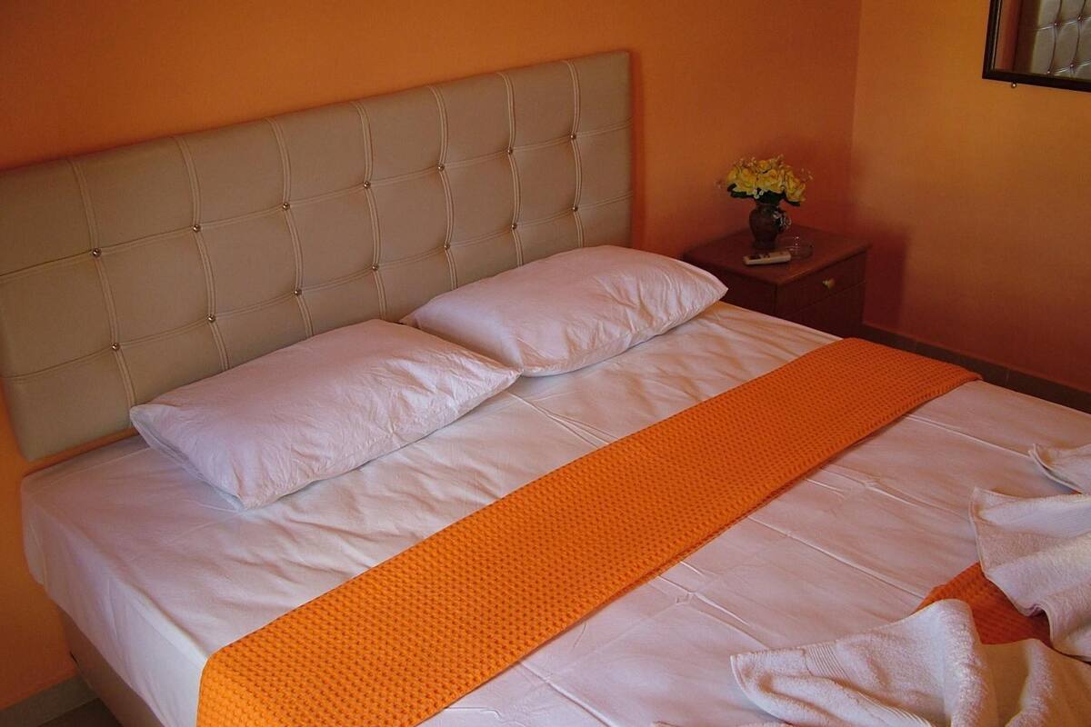 A neatly made bed with white linens and an orange throw blanket, a tufted headboard, and a small bedside table with a vase of flowers.