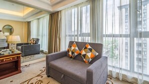 Una habitación de hotel con sofá, escritorio, lámpara y ventanas grandes con cortinas.