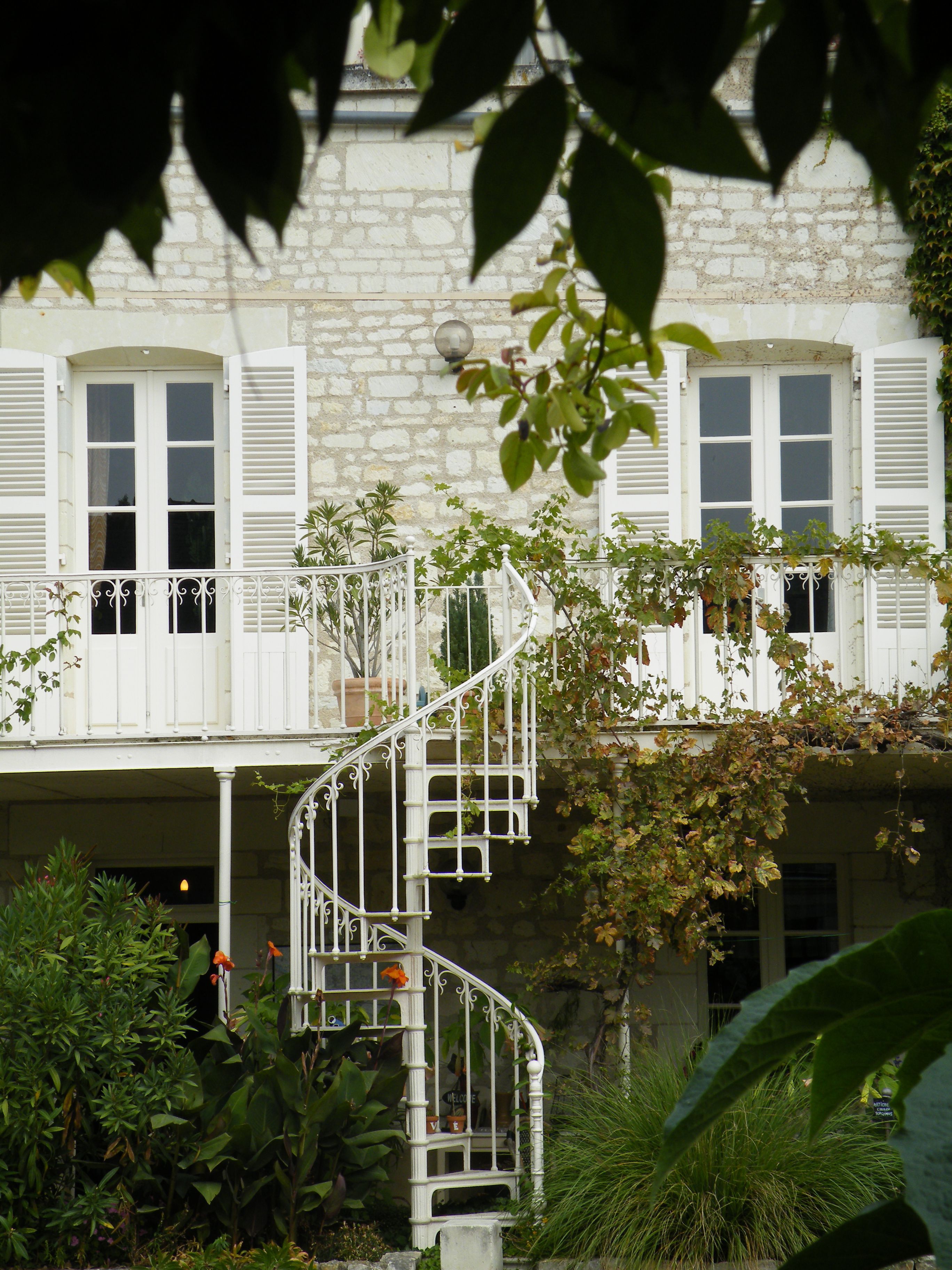 Un bâtiment en pierre avec des volets blancs, un escalier en colimaçon et une végétation luxuriante.