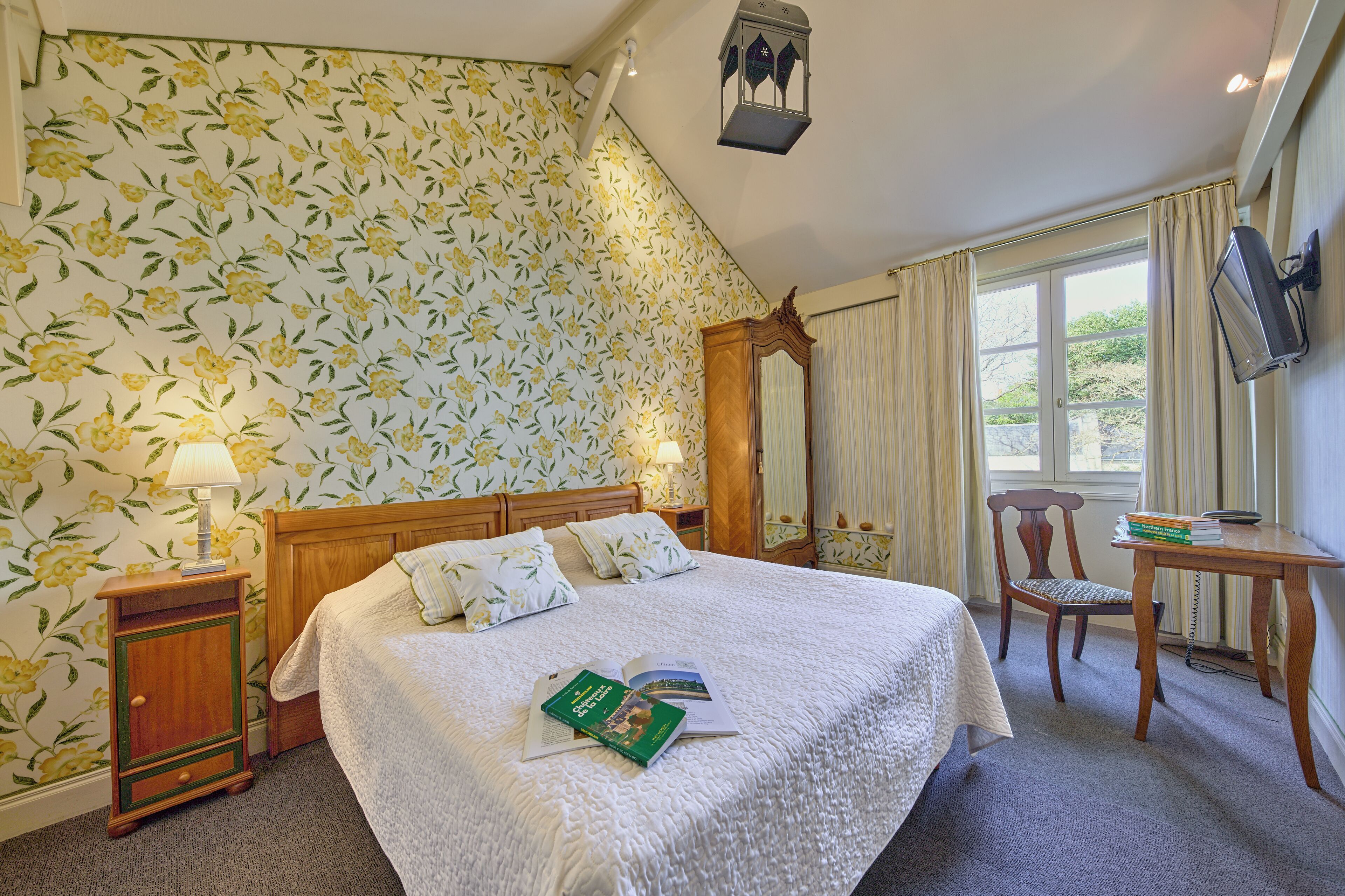 A neatly made bed with a floral wallpaper, a wooden nightstand with a lamp, a wooden chair, a small table with books, and a TV mounted on the wall.