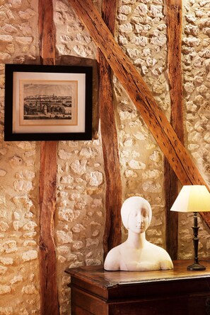 A rustic room with exposed wooden beams, a stone wall, a framed black and white photo, a bust on a wooden table, and a table lamp. - Hôtel Diderot (Chinon)