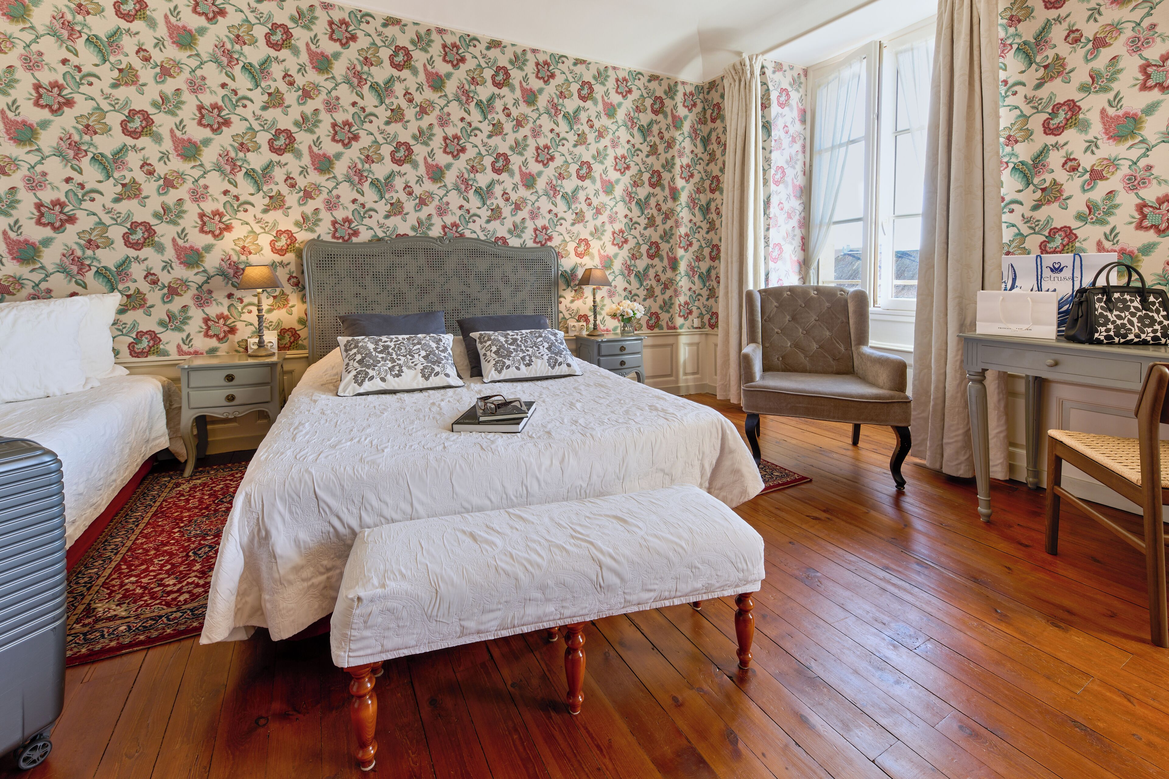 Quarto com papel de parede floral, uma cama grande com cabeceira de madeira, duas mesas de cabeceira com abajures, uma cadeira e uma escrivaninha com uma bolsa.