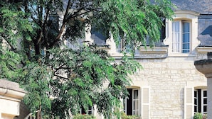 Un edificio de piedra con varias ventanas, un gran árbol en frente, y un patio de grava con una silla y plantas en macetas.