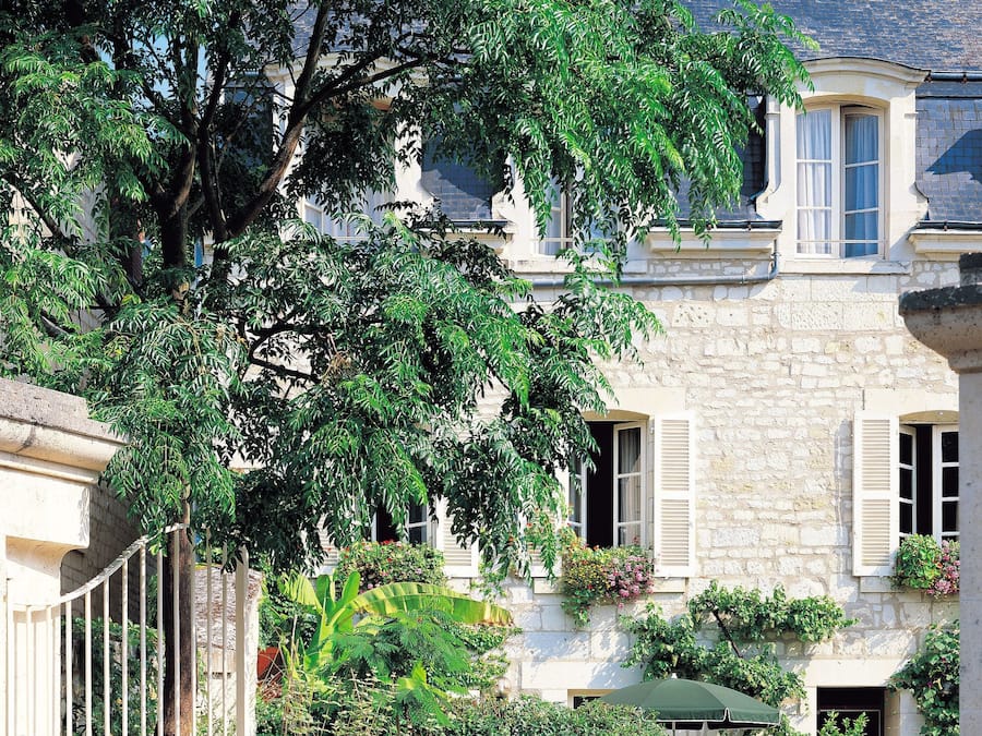 A stone building with multiple windows, a large tree in front, and a gravel courtyard with a chair and potted plants.