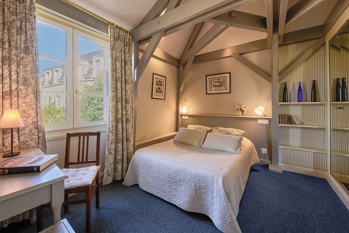 A bedroom with a bed, a desk, a chair, a window with curtains, and a bookshelf.