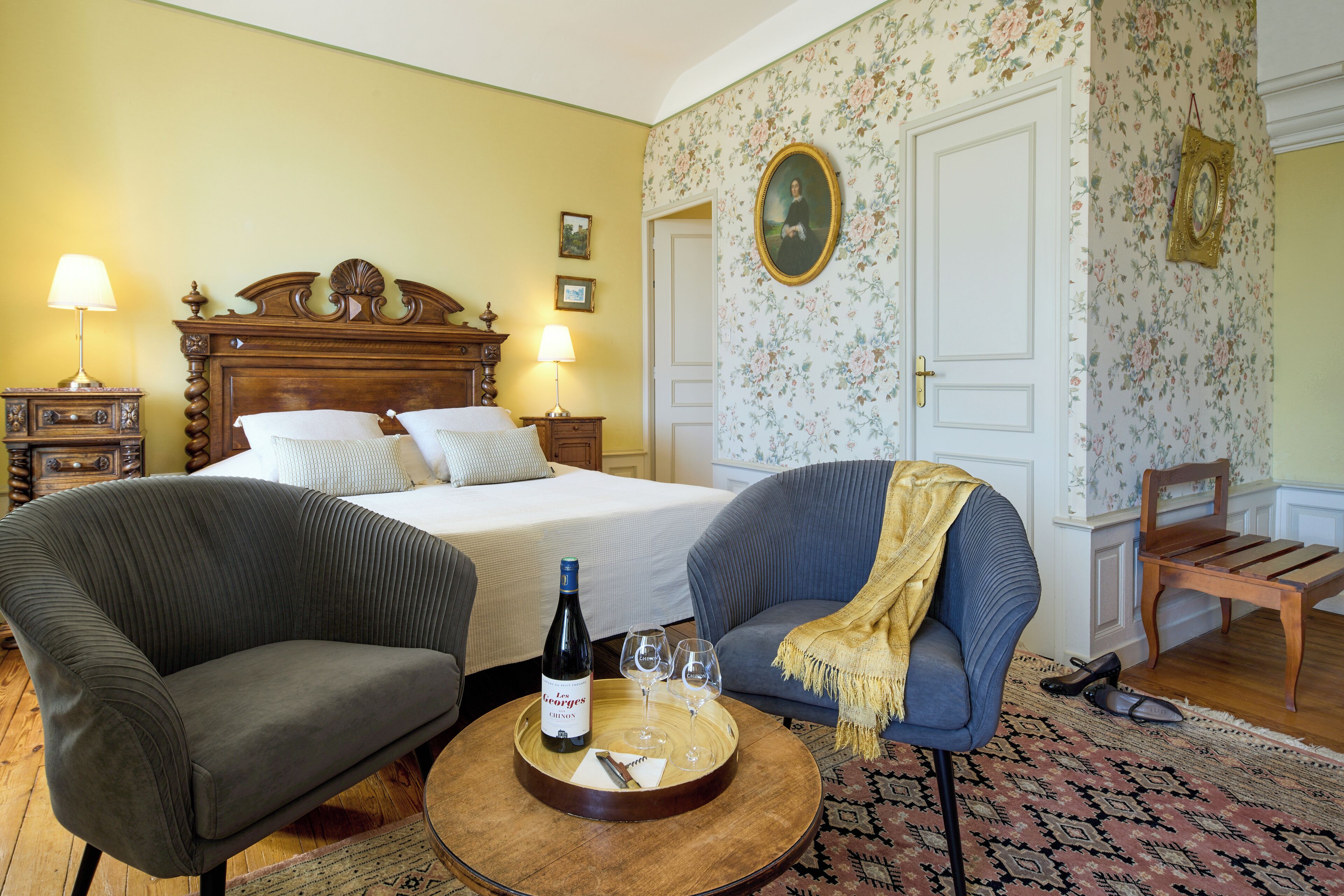 A hotel room with a bed, two armchairs, a wooden side table with a bottle and glasses, and a patterned rug.