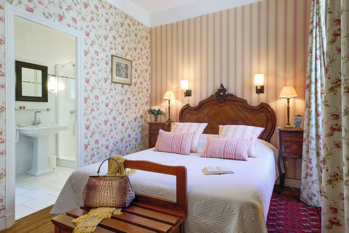 A hotel room with a bed, bedside lamps, a wooden headboard, a bedside table with a lamp, a mirror, a bathroom with a sink and toilet, and floral wallpaper.