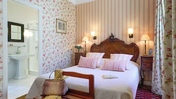 A hotel room with a bed, bedside lamps, a wooden headboard, a bedside table with a lamp, a mirror, a bathroom with a sink and toilet, and floral wallpaper.