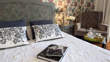 Een netjes opgemaakt bed met bloemenkussens, een nachtkastje met een lamp, een stoel en een dienblad met theepot en kopjes.