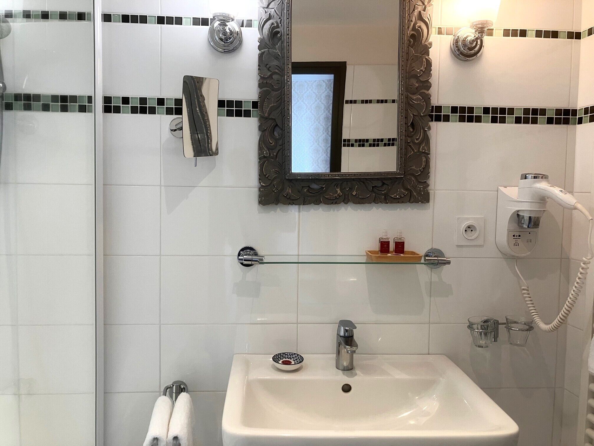 Une salle de bain avec un lavabo blanc, un miroir et une douche. On y trouve un porte-serviettes et une étagère en verre où sont rangés les articles de toilette.