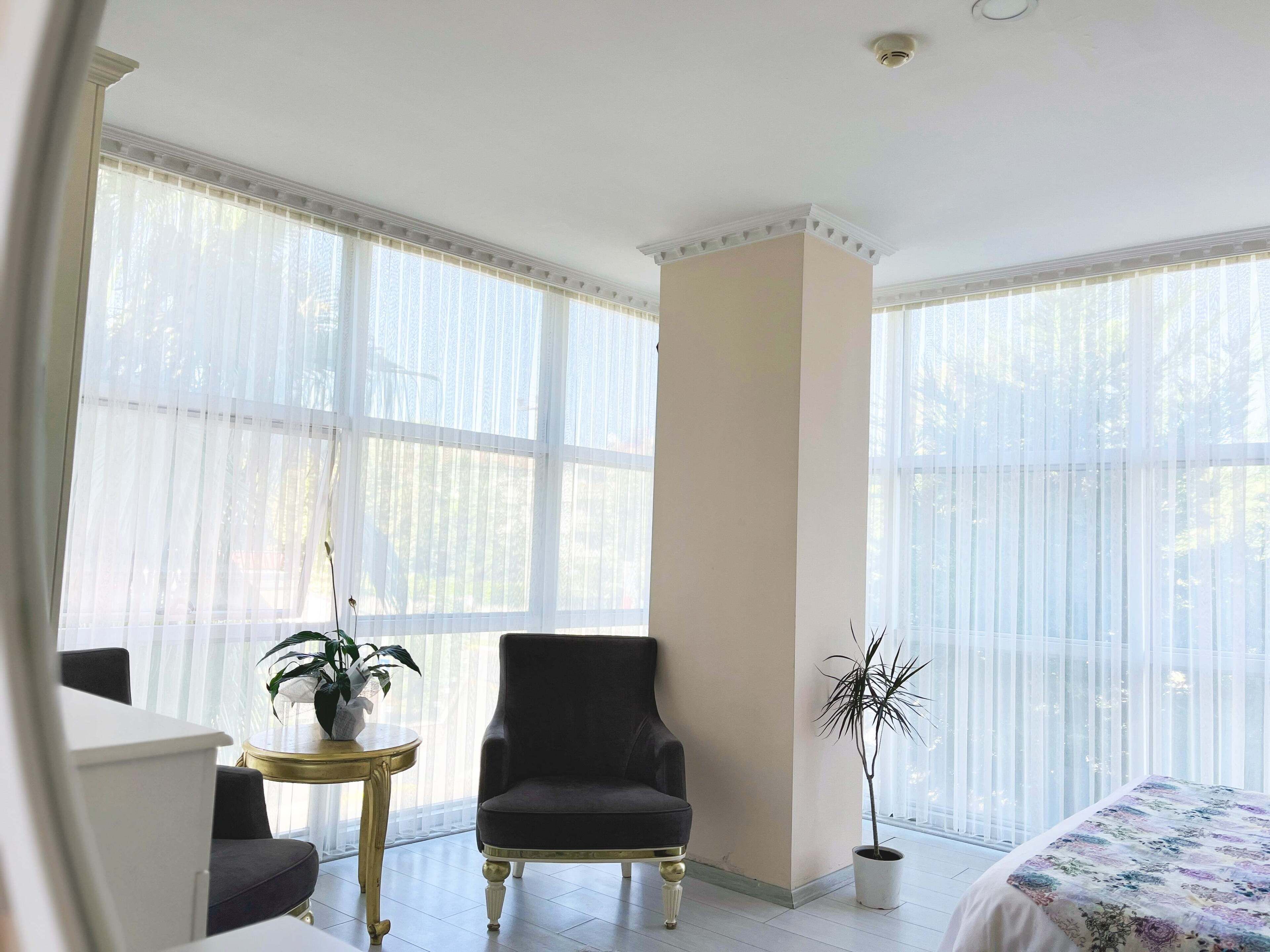A room with a bed, a dark armchair, a small round table with a plant, and a tall potted plant.