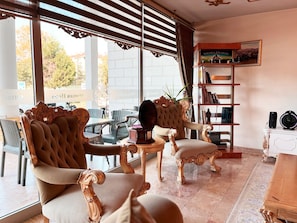 A room with two ornate chairs, a small table, a bookshelf, and a large window with blinds. - Ottoman Hotel Sakarya (Adapazari)