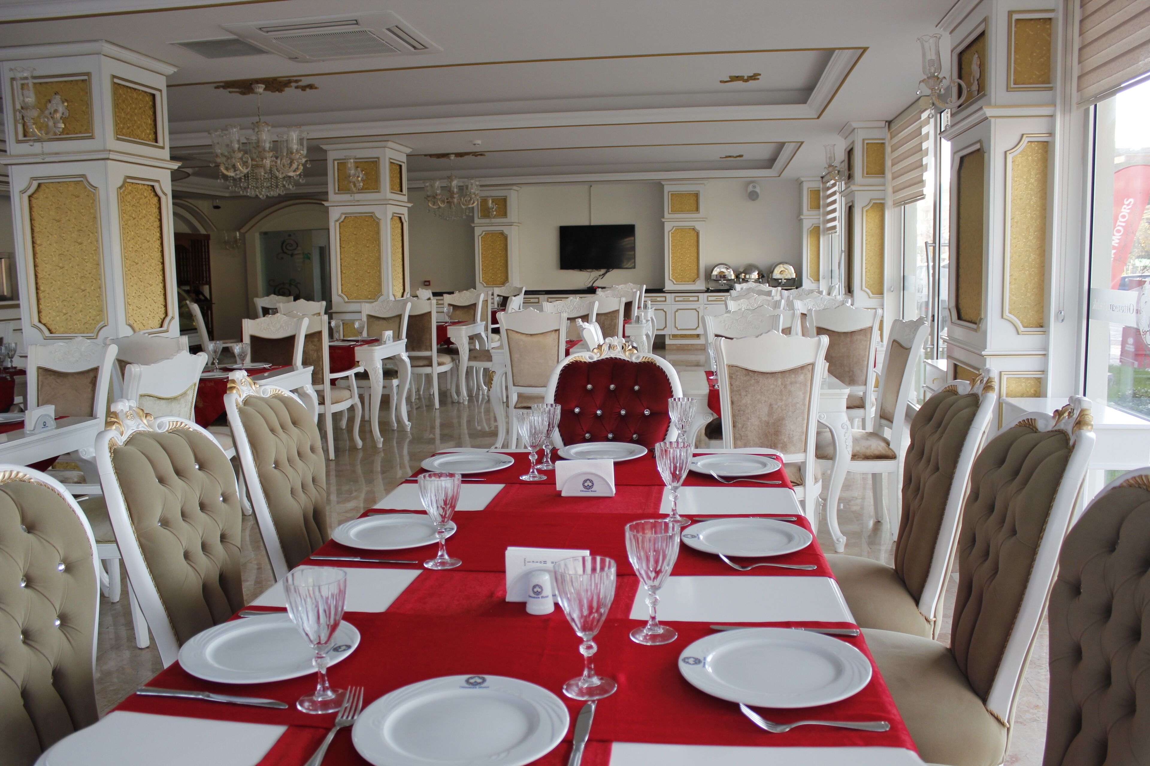 Un comedor formal con decoración blanca y dorada, varias mesas dispuestas para comer, y un televisor en la pared.