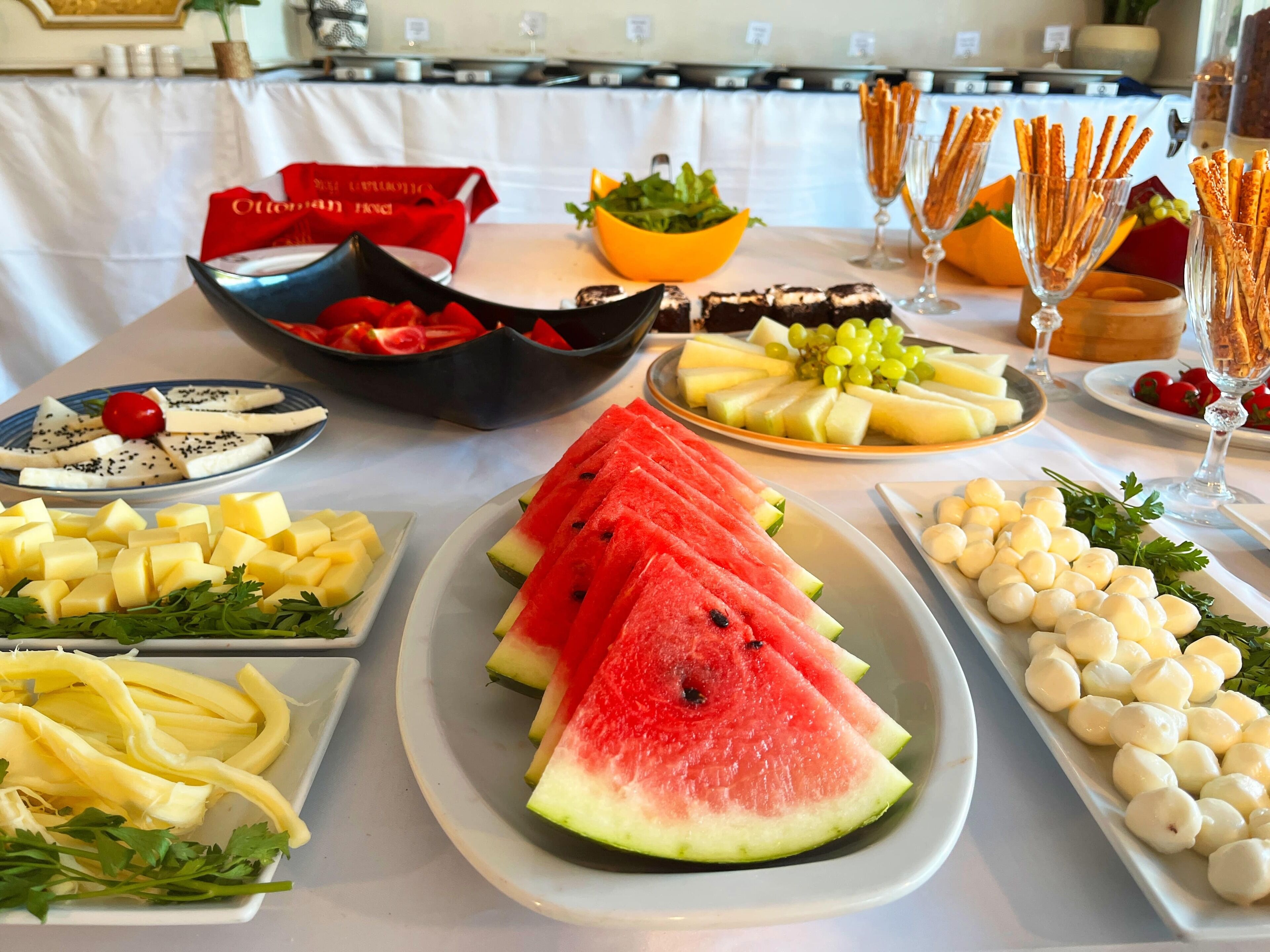A buffet table with various food items including watermelon slices, cheese, grapes, and breadsticks.