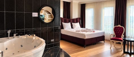 A modern hotel room with a large bathtub, a king-sized bed with a dark headboard, a red upholstered chair, and a round mirror on the wall.