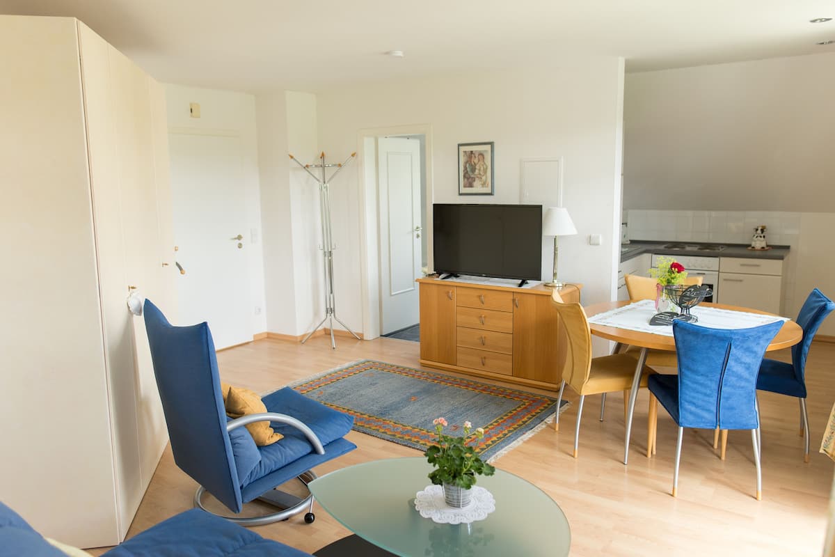 Un salon moderne comprenant un canapé, des fauteuils, une table basse en verre, un téléviseur et un petit coin cuisine.