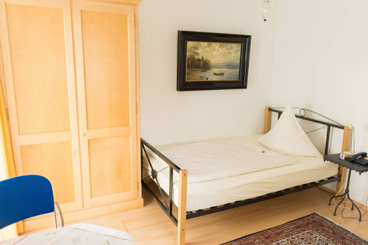 Une chambre avec un lit simple, une armoire en bois, une chaise, une table de chevet avec une lampe et un tableau au mur.