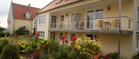 A two-story building with a balcony, a garden with various flowers, and a red-tiled roof.