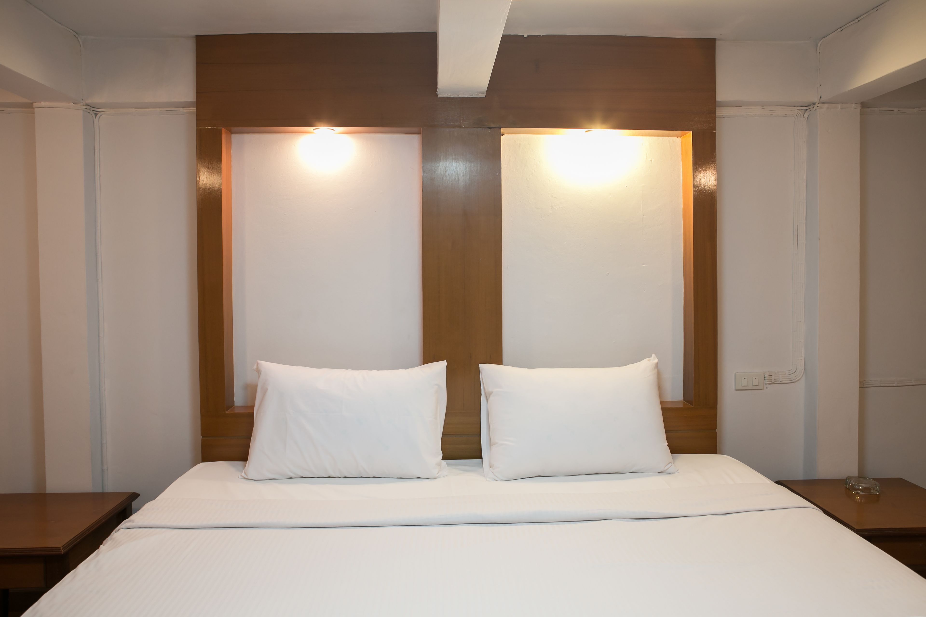 Habitación de hotel con una cama grande, dos almohadas blancas, cabecero de madera y dos luces fijadas a la pared.
