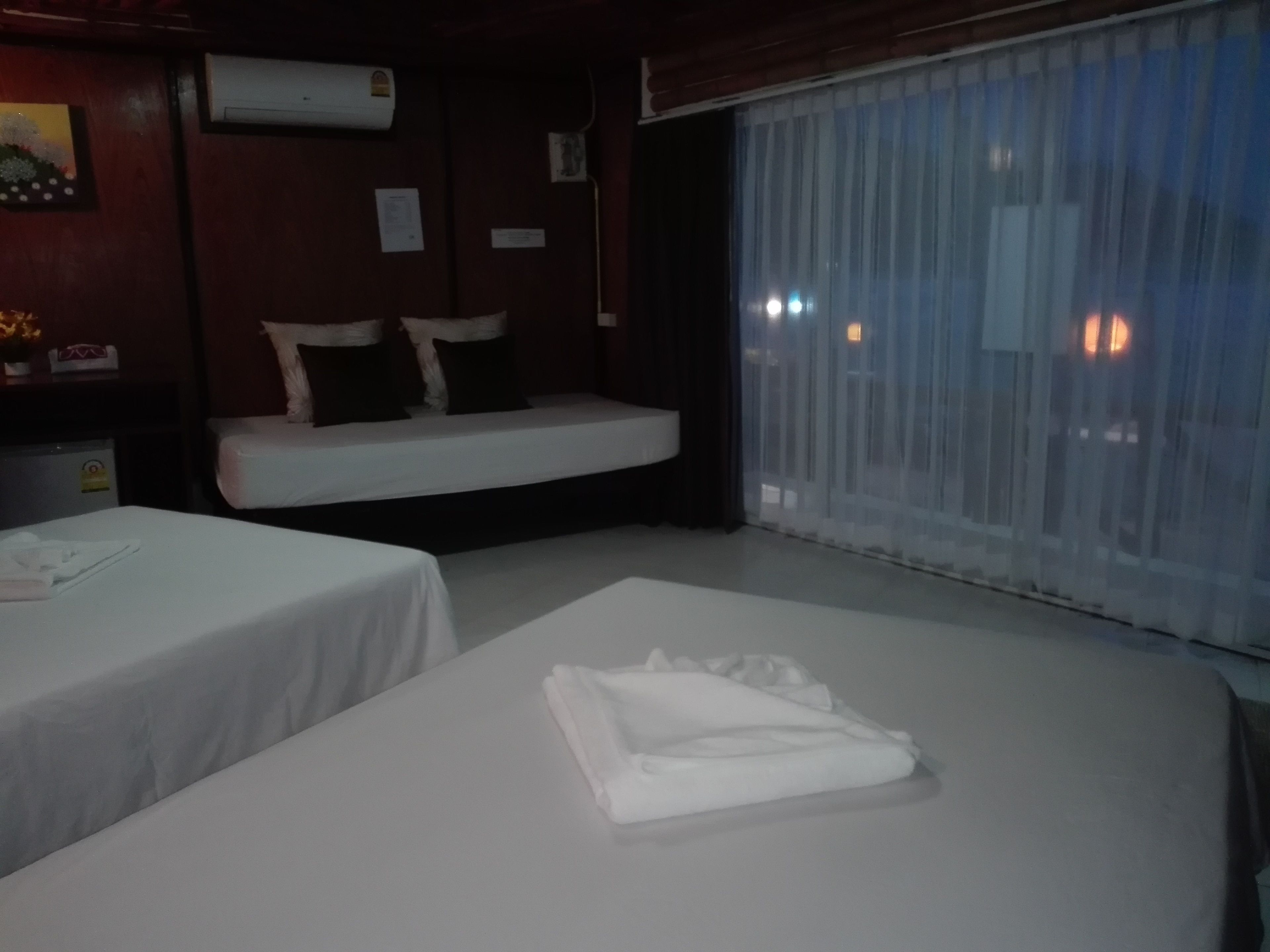 Habitación de hotel con dos camas, un sofá cama, una ventana con cortinas translúcidas y un equipo de aire acondicionado en la pared.