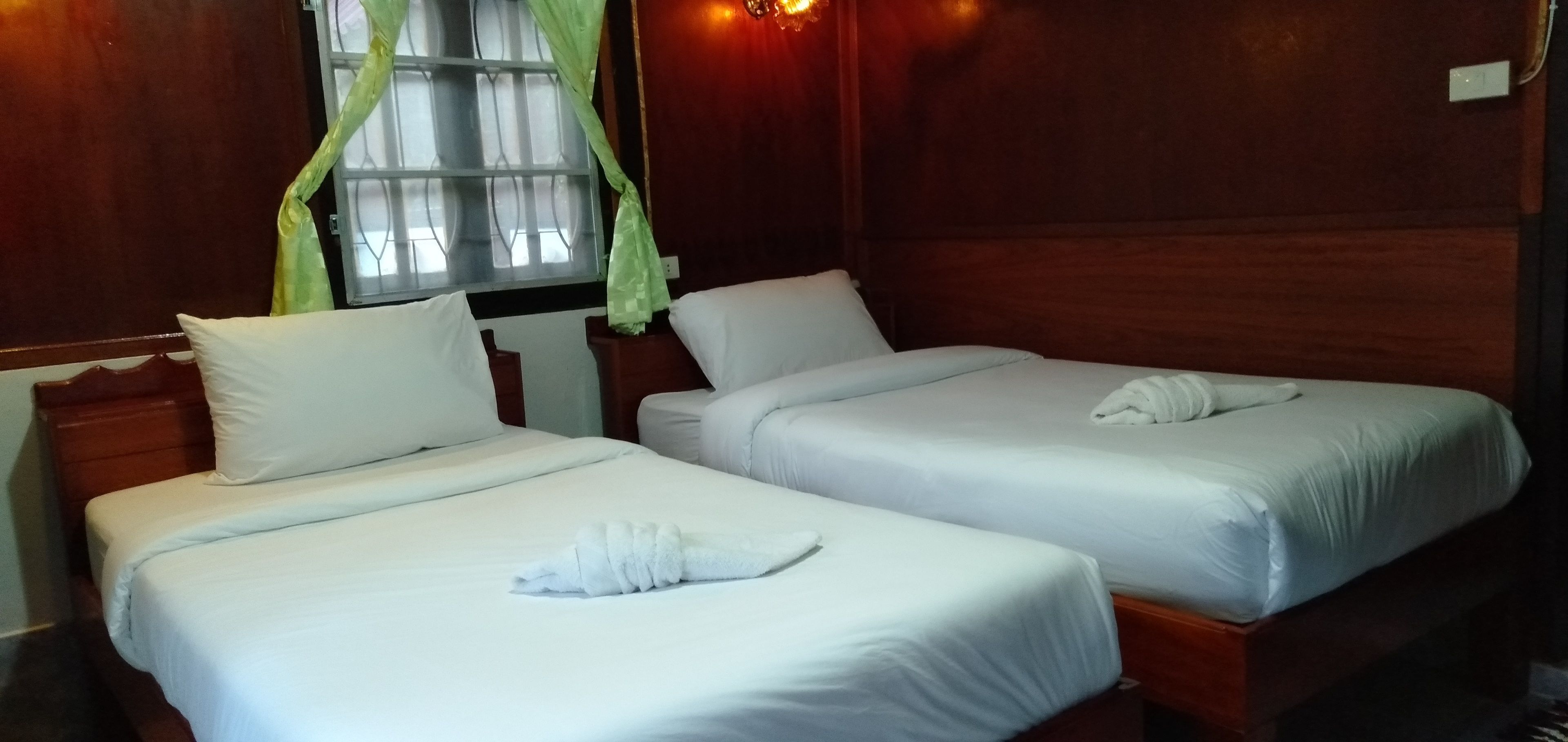 Two single beds with white linens and towels, a wooden headboard, a window with green curtains, and a wall-mounted light fixture.