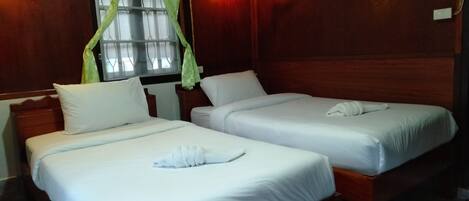 Two single beds with white linens and towels, a wooden headboard, a window with green curtains, and a wall-mounted light fixture.