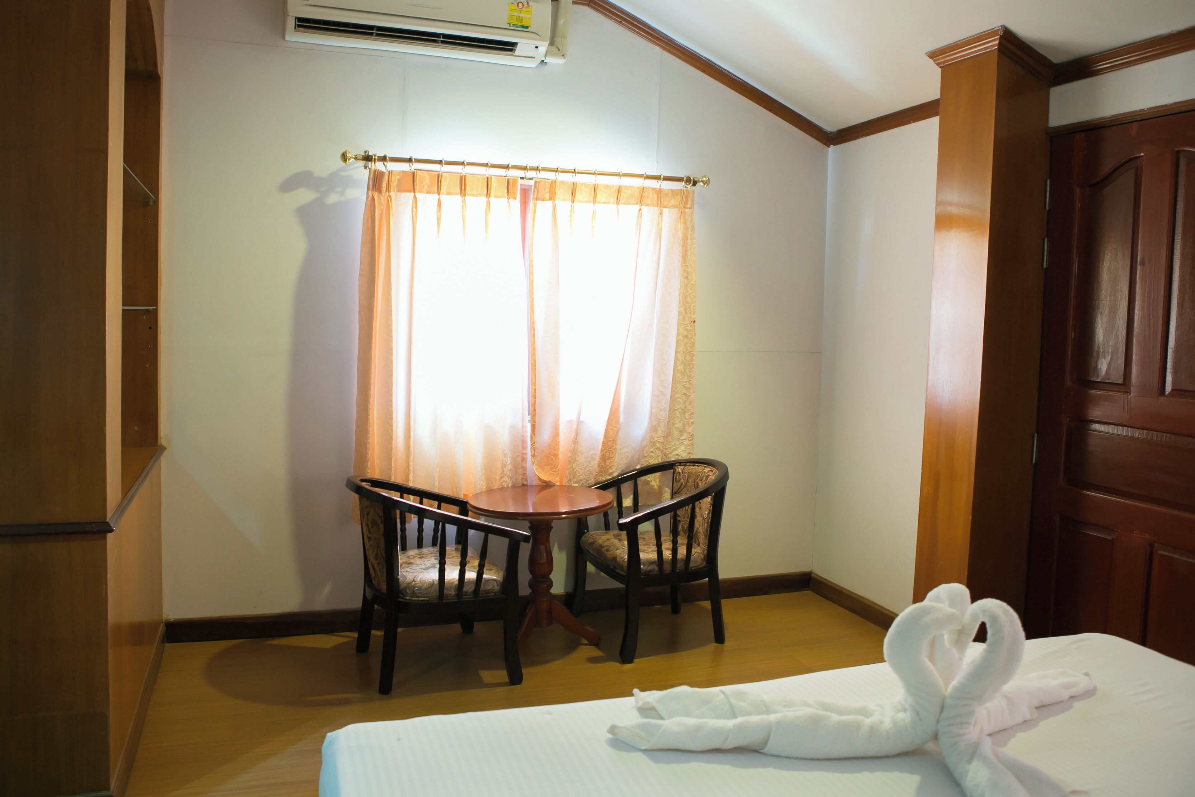 A hotel room with a bed, a wooden table with two chairs, a window with sheer curtains, and a wooden door.