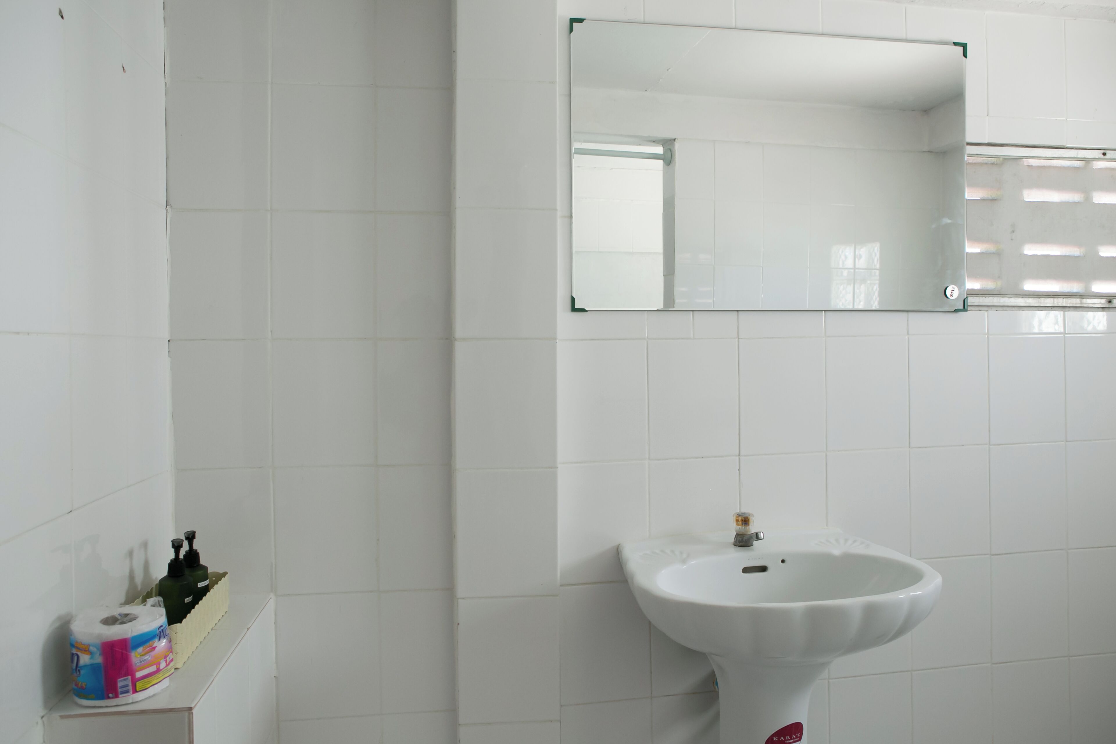 Une salle de bain avec des carreaux blancs, un lavabo blanc, un miroir et une étagère avec du papier toilette et une bouteille.