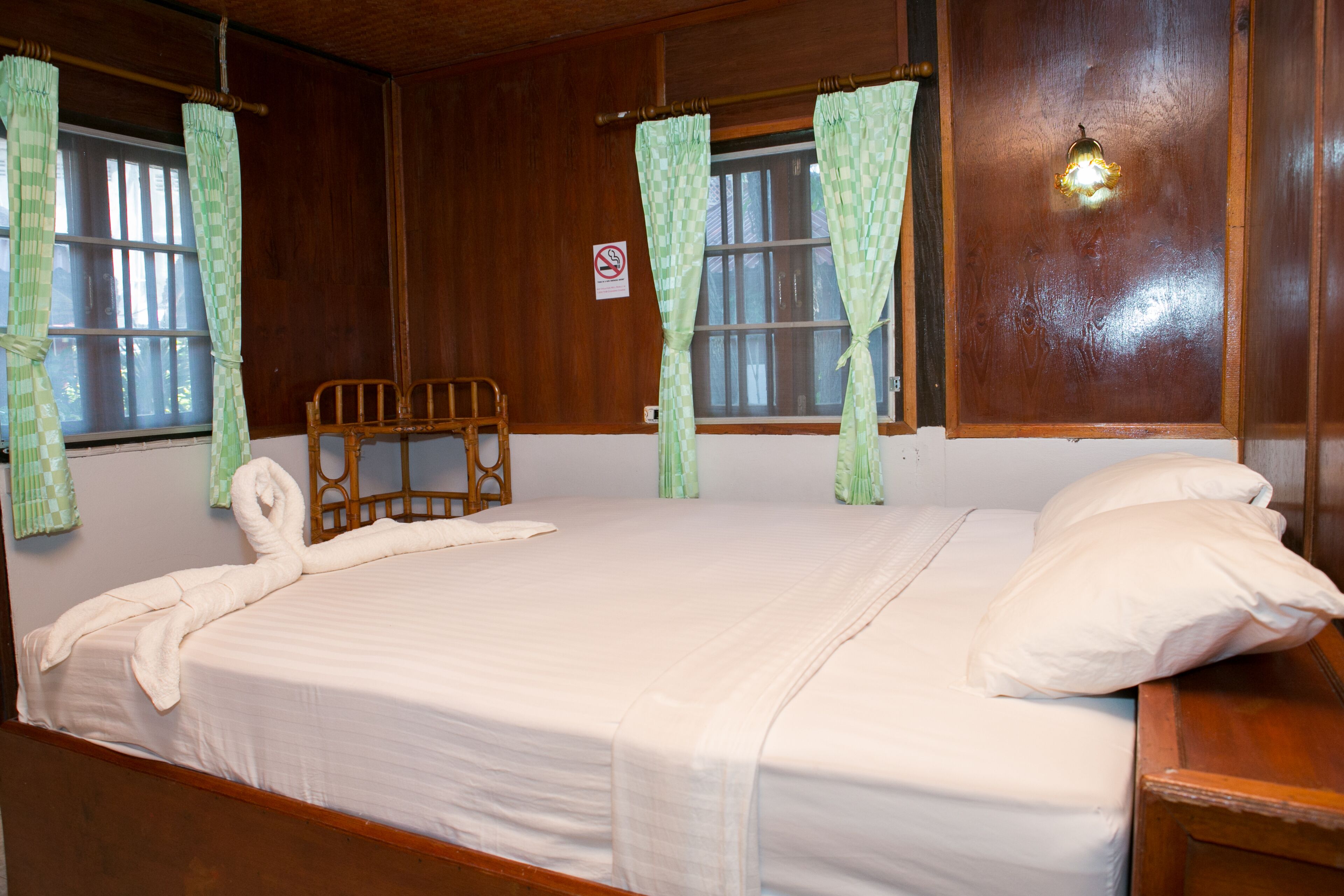A room with a bed, wooden headboard, two wooden chairs, a nightstand, a wall-mounted light, and a window with curtains.