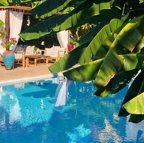 Zona de piscina con terraza de madera, sillas de descanso y un área de estar cubierta con vista a la piscina.