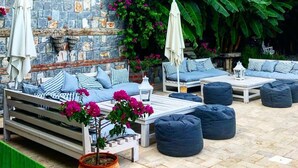 Un patio con pared de piedra, sofá blanco, cojines azules y una maceta con flores rosadas.