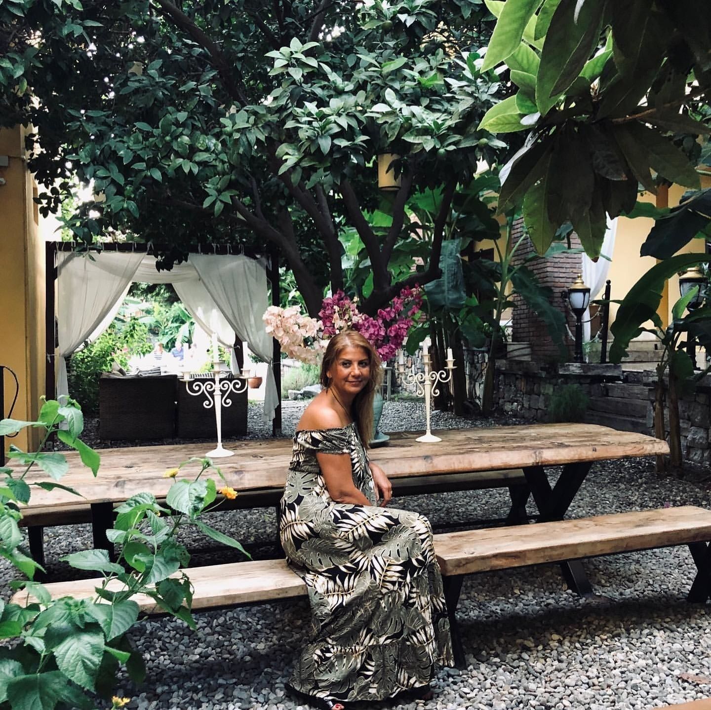 Una mujer sentada en un banco de madera en un jardín con un dosel blanco y un candelabro decorativo.