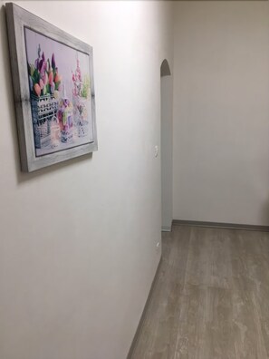 Käytävän vasemmalla seinällä on kehystetty taulu, lattia on puuta ja käytävän päässä on valkoinen ovi.