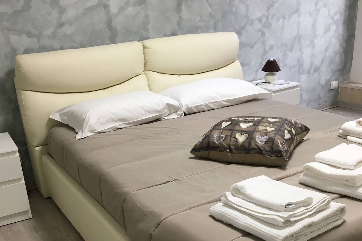 Een opgemaakt bed met witte en gemêleerde kussens, een gevouwen witte handdoek en een lamp op een nachtkastje.
