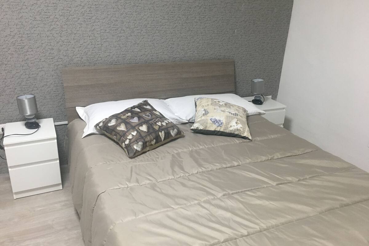 Een net opgemaakt bed met een beige dekbed, twee kussens met hartmotieven en een bedlampje op een witte nachtkastje.