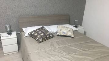 A neatly made bed with a beige comforter, two pillows with heart patterns, and a bedside lamp on a white nightstand.