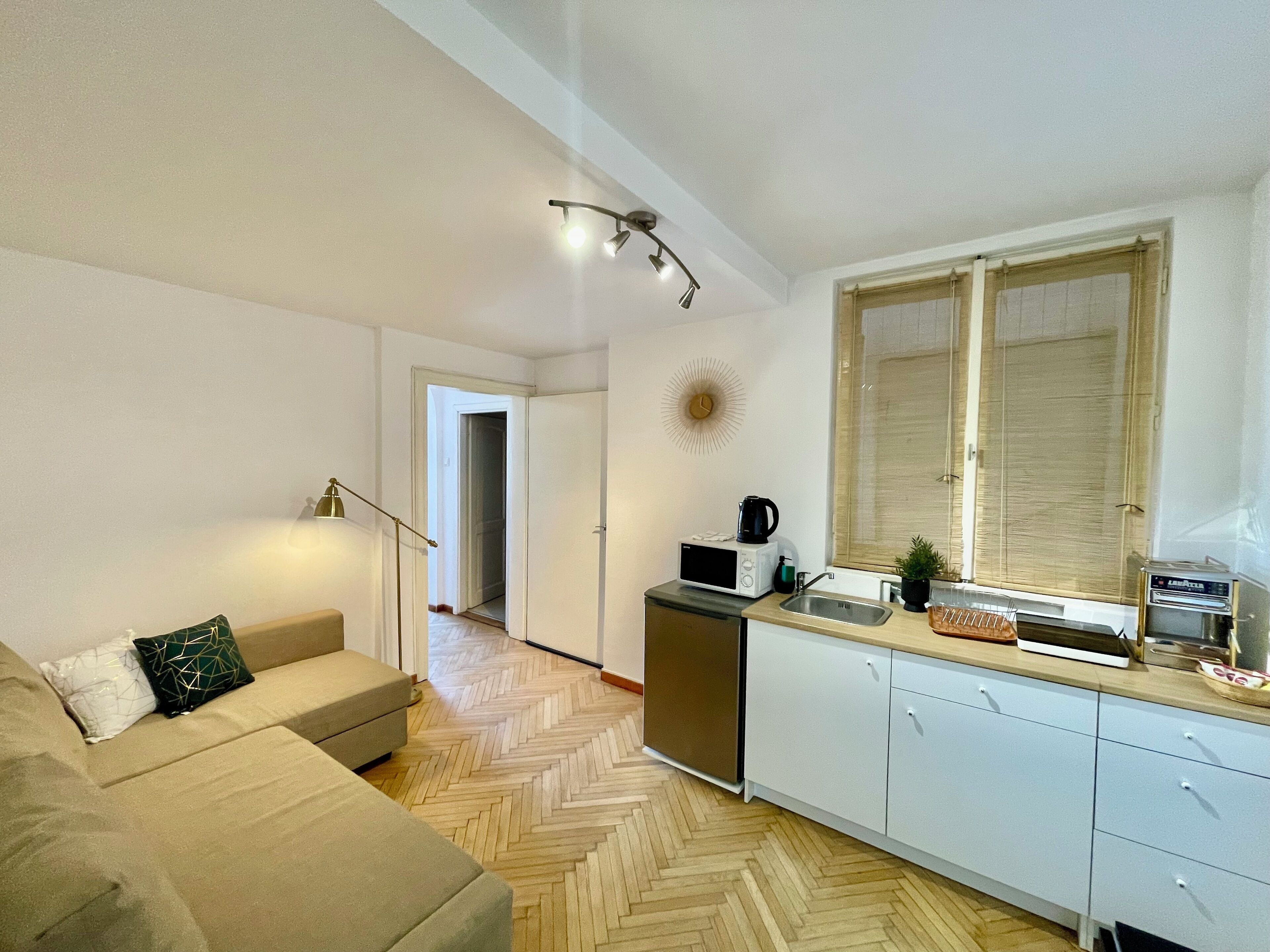 Una cocina moderna con microondas, cafetera y tostadora. El piso es de madera con patrón de espiga y tiene una ventana con persianas.
