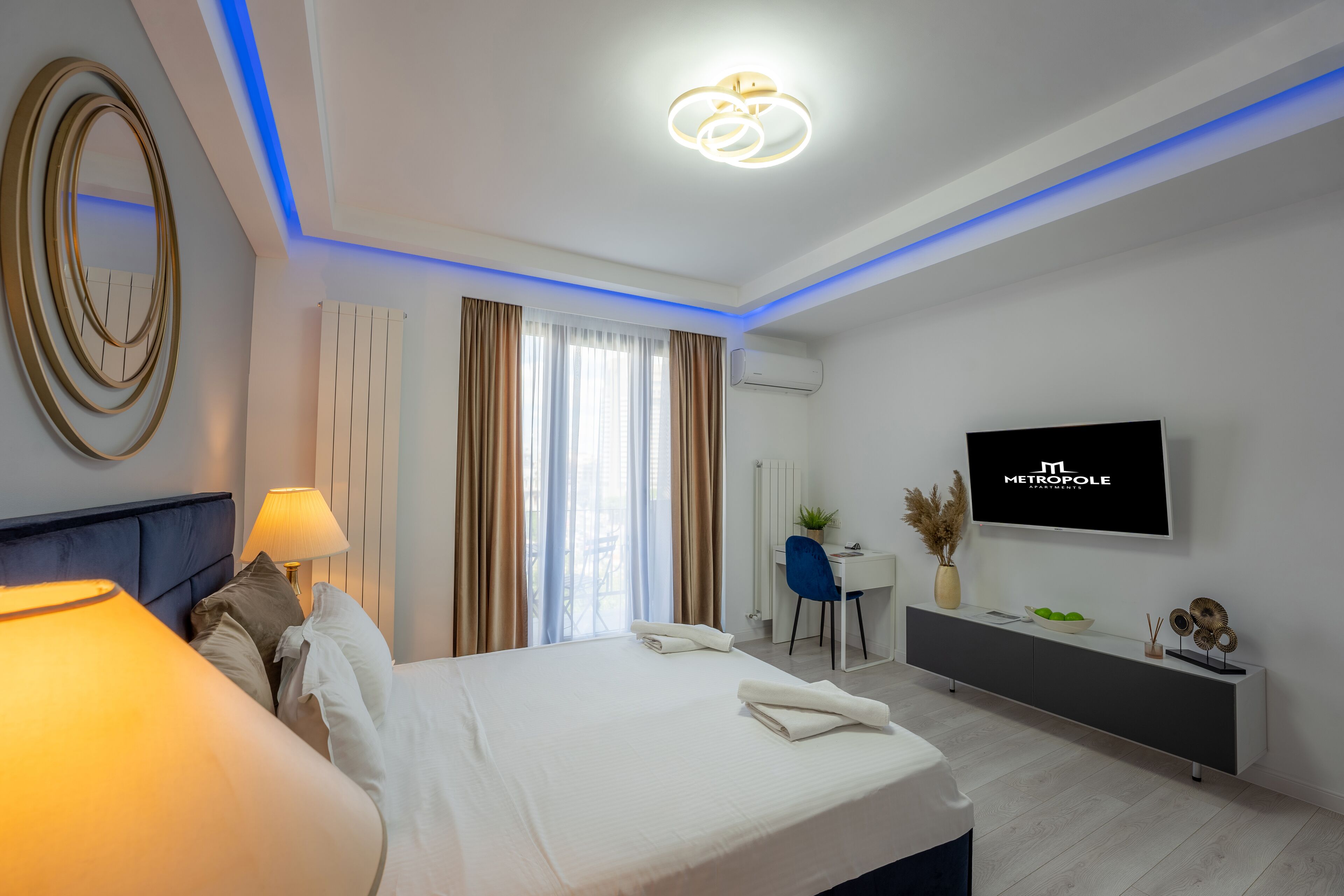 Quarto de hotel moderno com cama, abajur, televisão, escrivaninha e vista para o exterior.