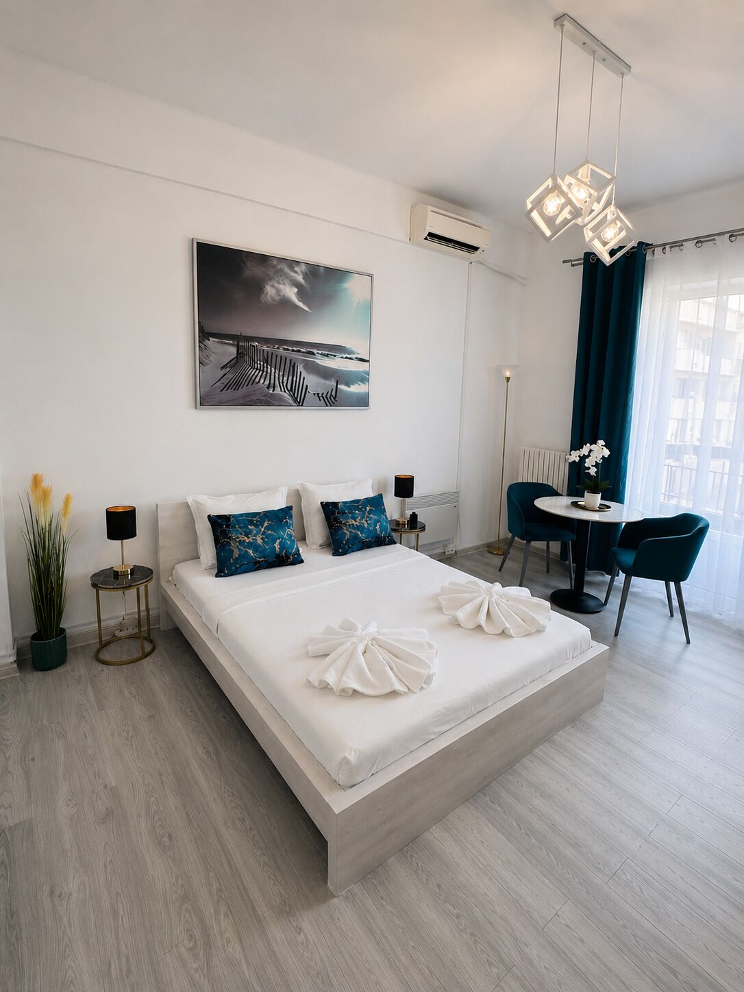 A modern hotel room with a large bed, a painting on the wall, a desk with chairs, and a view of the outdoors.