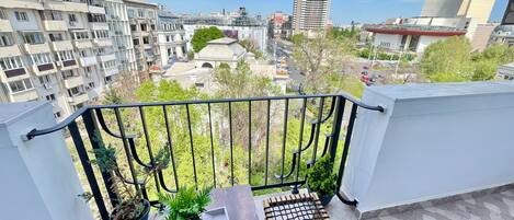 Un balcón con mesa y sillas, plantas en macetas y vistas de edificios circundantes.
