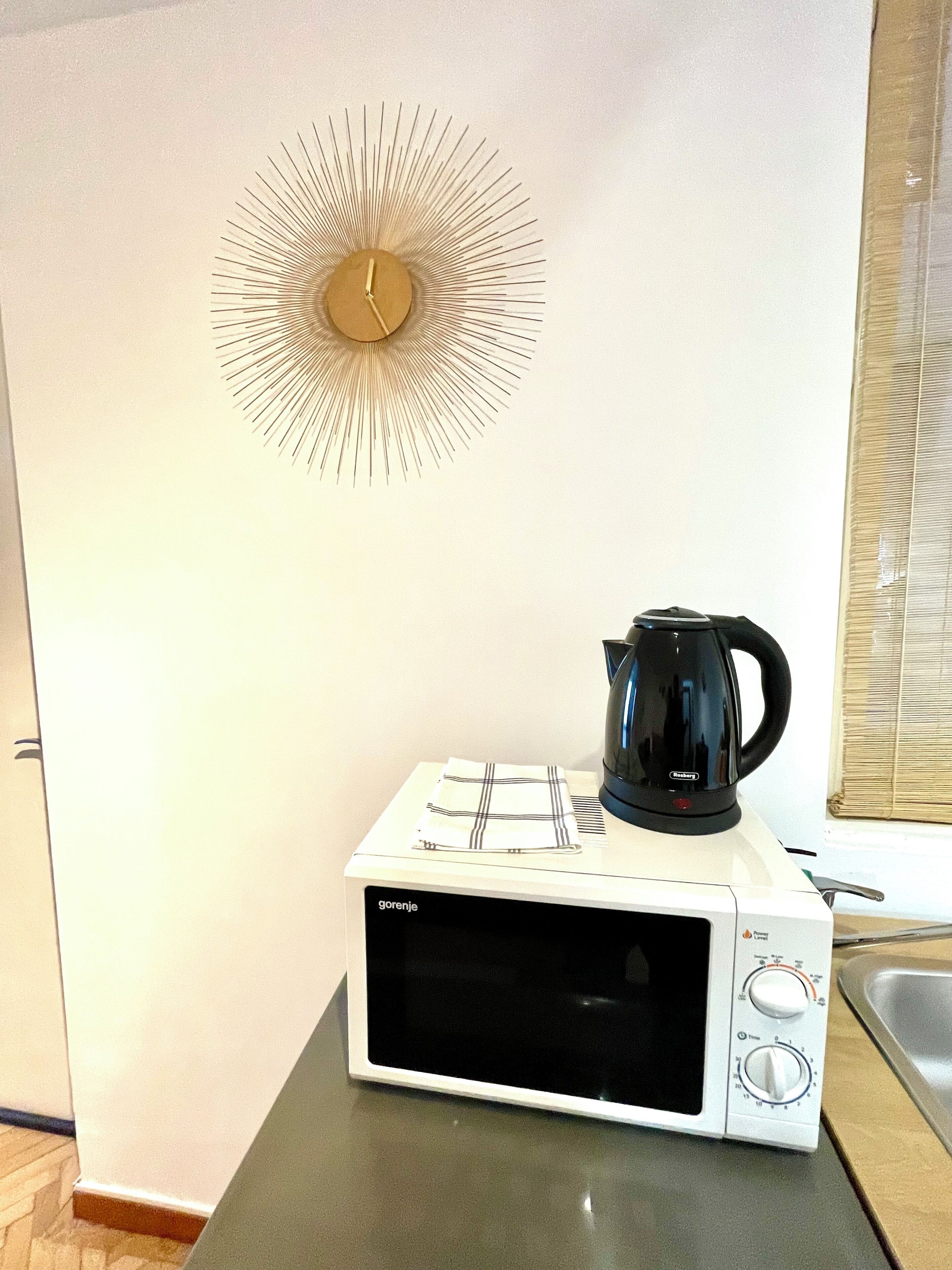 A white microwave oven with a black coffee pot on top, a wall clock, and a window with blinds.