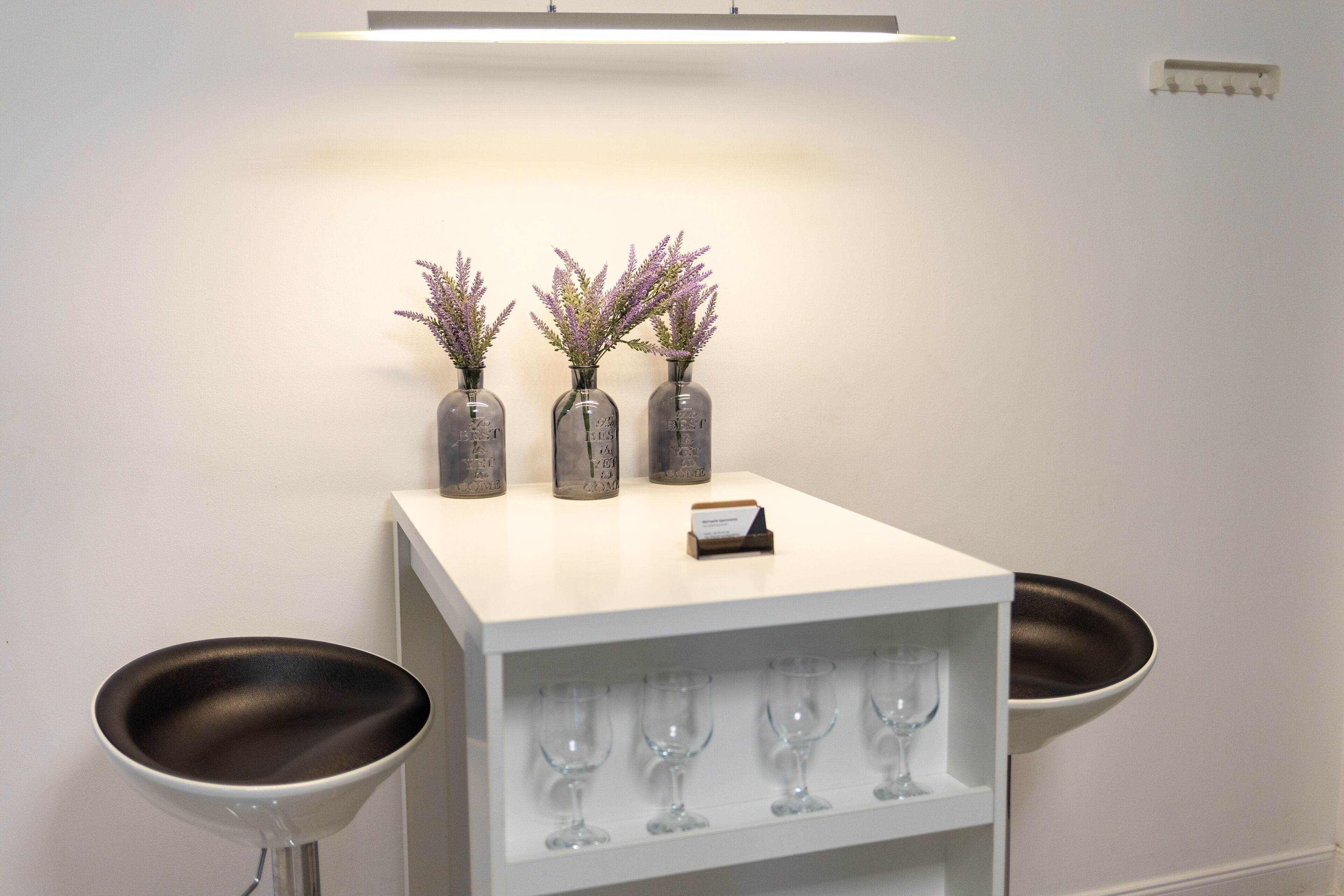 A white bar area with three vases of purple flowers, a small card, and a shelf with wine glasses.