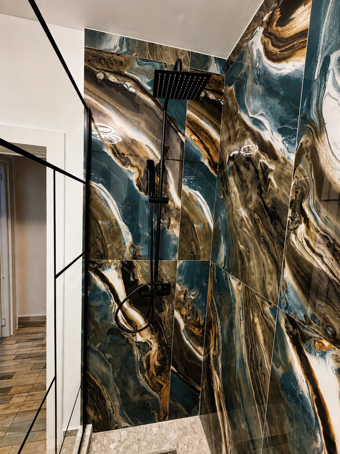 Une salle de bain avec un espace douche, un mur carrelé orné d’un motif évoquant le marbre, et une porte visible donnant sur une autre pièce.