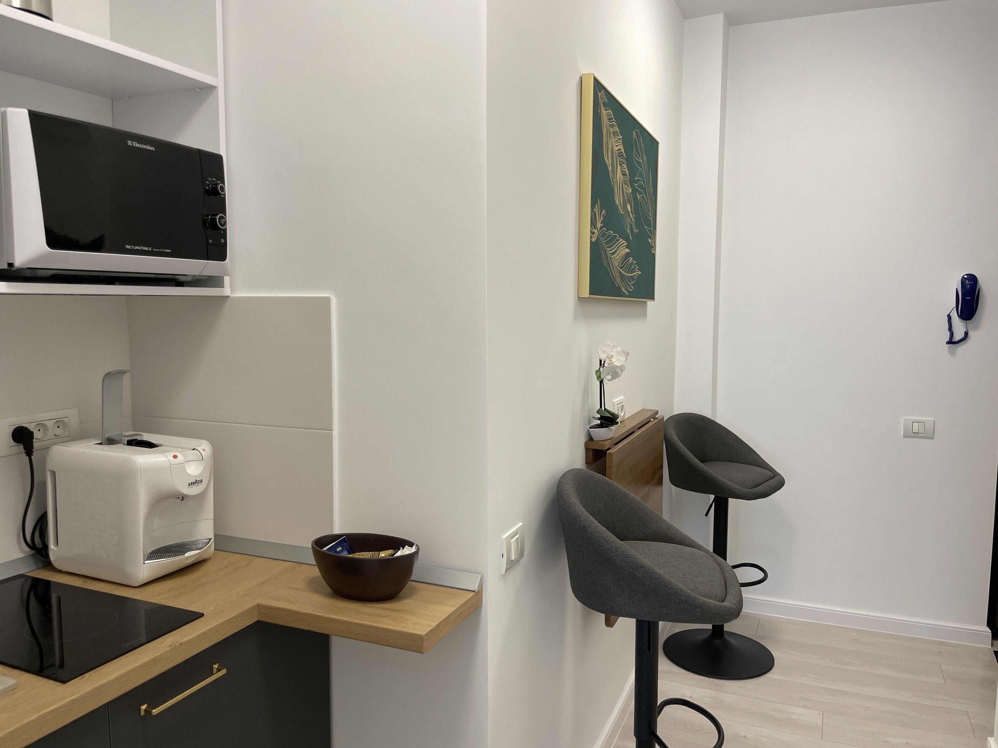 Une cuisine moderne équipée d’un four à micro-ondes, d’un grille-pain et d’un bol posé sur une étagère en bois. On y trouve deux chaises grises et une petite table sur laquelle est placée une orchidée.