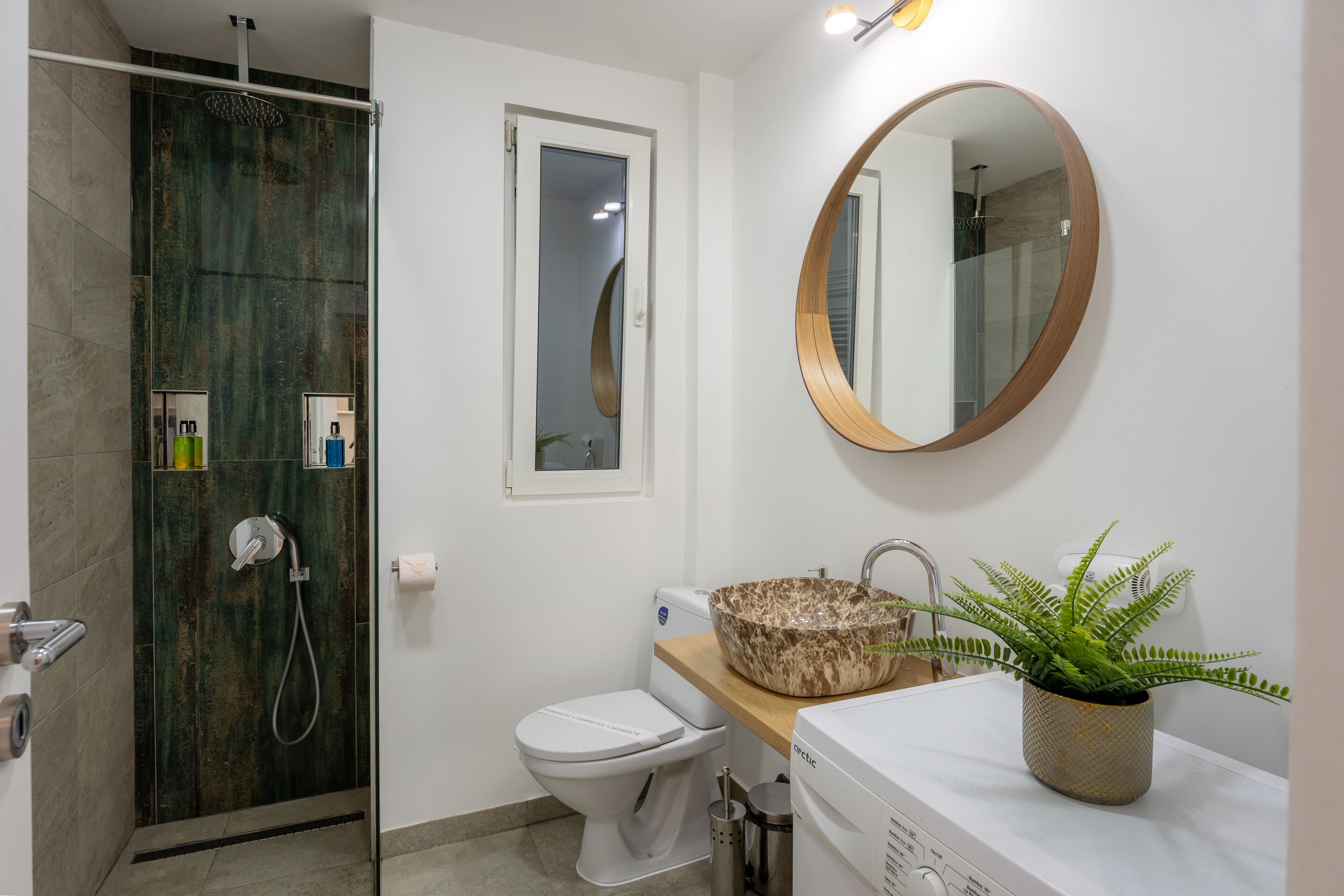 A bathroom with a glass-enclosed shower, a toilet, a sink with a marble basin, and a round mirror on the wall.