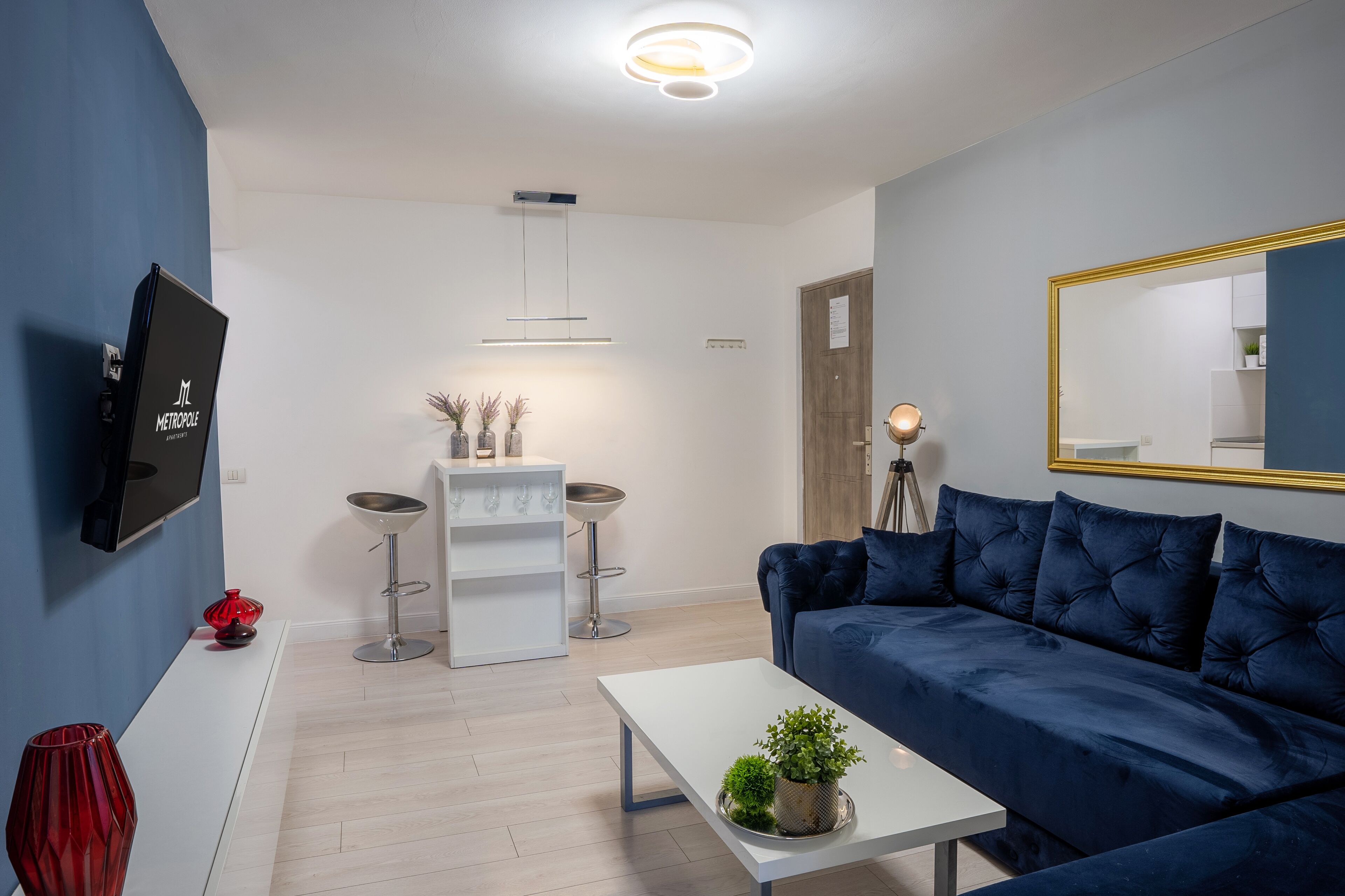 A modern living room with a blue sofa, white coffee table, and a TV mounted on the wall. There are two bar stools and a small white cabinet with decorative items.
