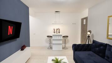 A modern living room with a dark blue sofa, a white coffee table with a plant, a TV mounted on the wall, and a minimalist side table with two stools.
