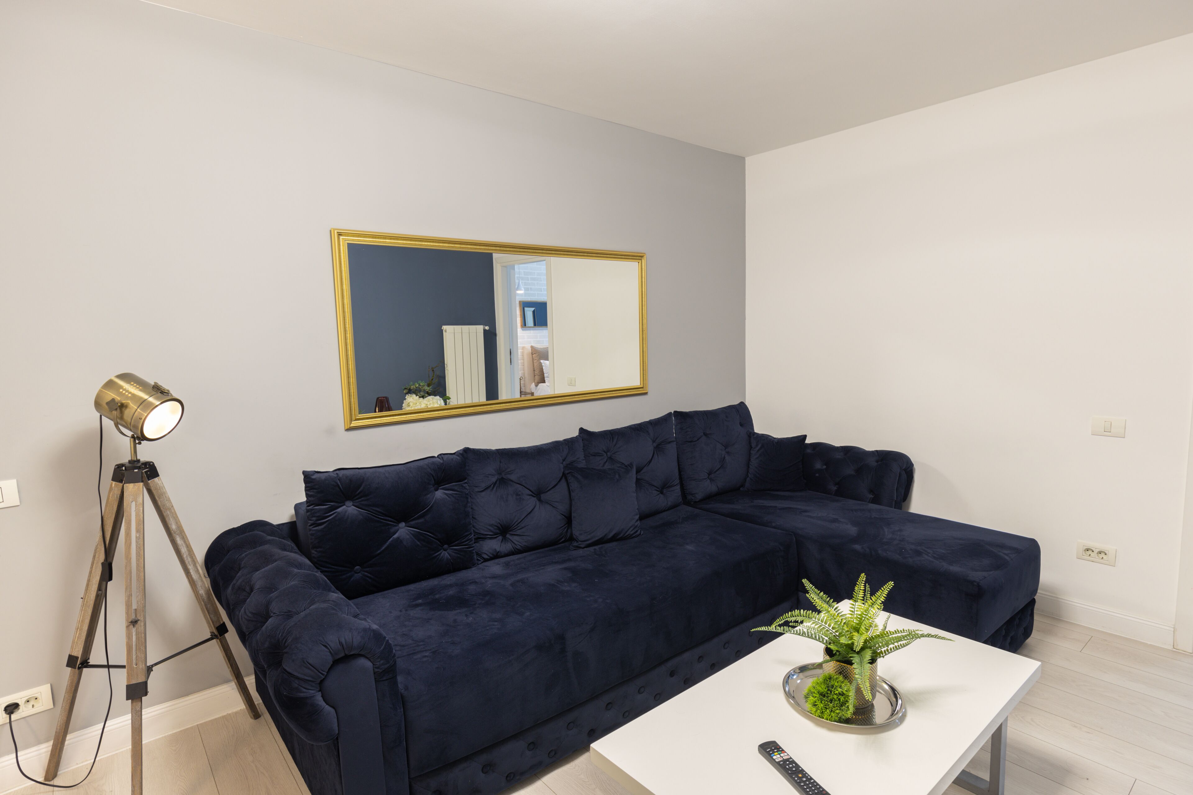 A modern living room with a dark blue sofa, a white coffee table with a plant, a tripod with a light, and a large mirror on the wall.
